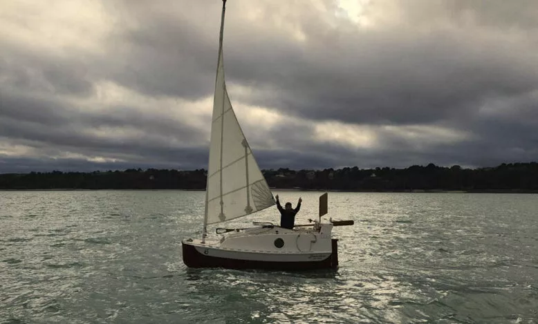Une personne sur un petit voilier les bras levés, en mer sous un ciel nuageux