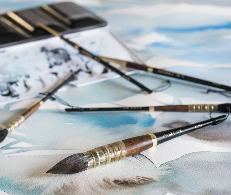 Pinceaux Isabey pour aquarelle posés sur une palette d’artiste avec lavis bleu en fond