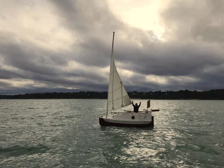 Solo sailor raising arms in triumph on a sailboat sponsored by Raphaël on open water