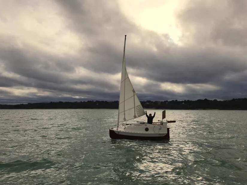 Solo sailor raising arms in triumph on a sailboat sponsored by Raphaël on open water