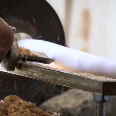 Close-up of artisan shaping a wooden brush handle on a lathe with wood shavings flying